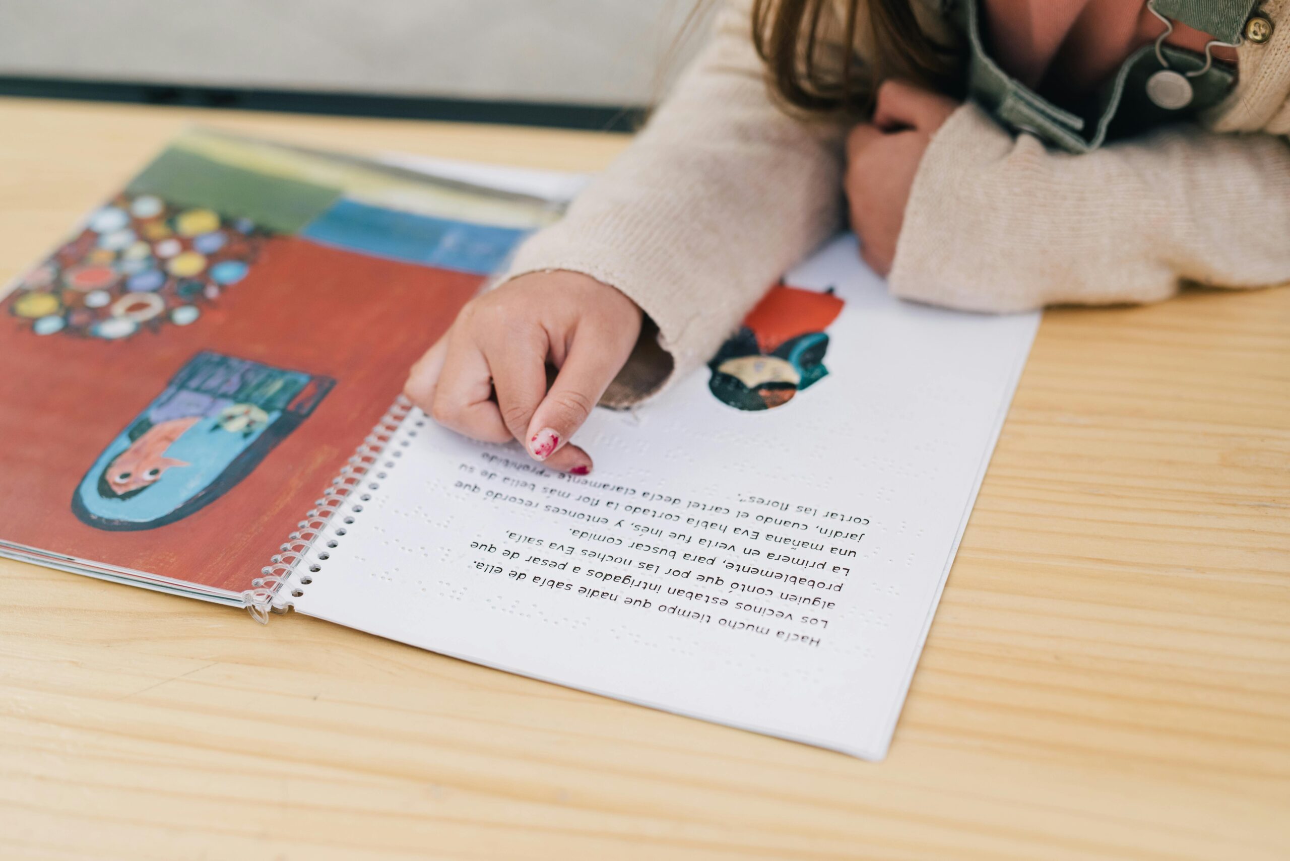 Child's hand pointing at illustrations in a colorful book on a wooden table.